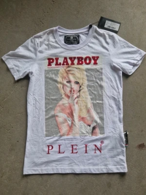 Vit Playboy t-shirt från Philipp Plein - Vit t-shirt från Philipp Plein med stort Playboy-tryck framtill och röda detaljer. Klassisk rund halsringning och korta ärmar. T-shirten har en modern och edgy vibe, perfekt för dig som vill sticka ut med en statement-piece.                         Kan eventuellt mötas upp i Stockholm för ett billigare pris!!!