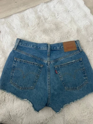 Levi's blå denimshorts med slitningar - Snygga blå denimshorts från Levi's med hög midja och råa kanter. Shortsen har slitna detaljer framtill och klassiska bakfickor med Levi's-logga. Perfekta för sommaren och ger en cool, avslappnad vibe.