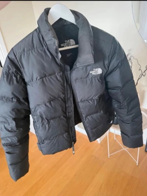 Svart pufferjacka från The North Face - Snygg svart pufferjacka från The North Face med hög krage och dragkedja framtill. Jackan har en klassisk boxig passform och tydliga quiltade sömmar. Perfekt för kalla dagar och riktigt varm tack vare sitt vadderade material.