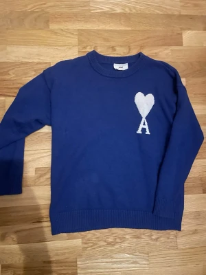 Blå stickad tröja från Ami - Snygg blå stickad tröja från Ami Alexandre Mattiussi med ett vitt hjärta och bokstaven A på bröstet. Tröjan har rund halsringning och ribbade muddar vid ärmslut och nederkant. Perfekt för dig som gillar stilrena detaljer.