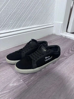 Svarta Saint Laurent sneakers - Säljer ett par svarta sneakers från Saint Laurent med vit sula och handskriven logga på sidan. Skorna har lågt skaft, svarta snören och ovandel i tyg och läder. Perfekta för dig som gillar stilrena och klassiska sneakers med en twist.