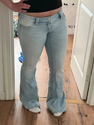 Ljusblå bootcut lågmidjade jeans - Säljer ett par ljusblå bootcut jeans med snygga pärldetaljer på fickorna. Jeansen har klassisk femficksdesign och är tillverkade i denim med stretch. Perfekt för dig som gillar en retrostil och vill ha ett par jeans som sticker ut.
