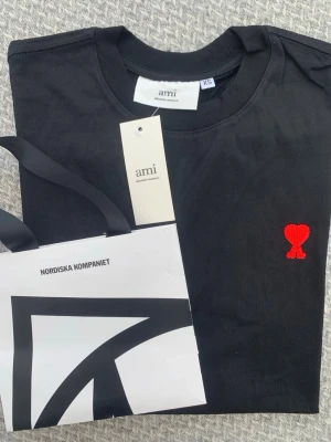 Svart t-shirt från Ami med rött hjärta - Svart t-shirt från Ami Alexandre Mattiussi med klassisk passform och rund hals. På bröstet finns ett broderat rött hjärta. T-shirten är i mjuk bomull och har korta ärmar. Perfekt för en stilren och trendig look.