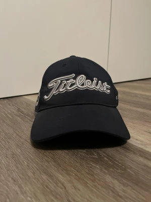 Mörkblå Titleist keps med brodyr - Snygg svart keps från Titleist med broderad logga framtill och texten 'Pro V1' och ’FJ’ på sidan. Klassisk böjd skärm och justerbar passform. Perfekt accessoar för golfbanan eller en sportig look. Pris kan absolut diskuteras.     