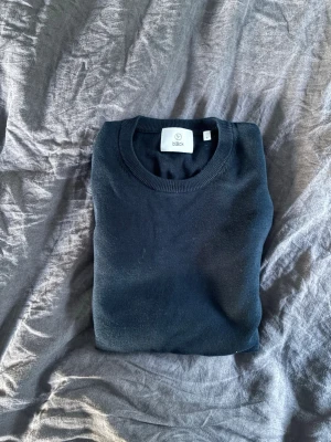 Svart snygg tröja från Bläck - En stilren svart tröja från Bläck med rund halsringning och långa ärmar. Tröjan har ribbade muddar vid ärmslut och nederkant, perfekt för en clean och enkel look. Passar dig som gillar minimalistisk stil.