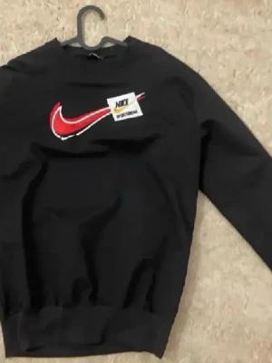 Svart Nike sweatshirt med röd swoosh - Svart sweatshirt från Nike med en stor röd och vit swoosh-logga på bröstet samt en Nike Sportwear-patch. Tröjan har rund halsringning och ribbade muddar vid ärmslut och midja. Perfekt för en sportig och avslappnad stil.