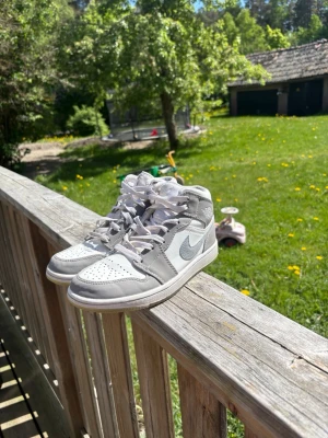 Nike Air Jordan 1 Mid grå/vit - Snygga Nike Air Jordan 1 Mid sneakers i grått och vitt med klassisk siluett. Skorna har vit tåbox, gråa paneler och vit swoosh på sidan. Hög modell med snörning och ikonisk Air Jordan-logga vid ankeln. Perfekta för dig som gillar streetwear och sportig stil.