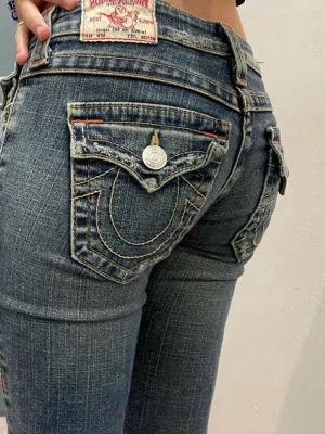 True Religion bootcut jeans blå denim - Säljer ett par blå bootcut jeans från True Religion med ikoniska kontrastsömmar och detaljerade bakfickor med lock och silverfärgade knappar. Jeansen har låg midja och klassisk femficksdesign. Materialet är denim i bomull med lätt sliten look.