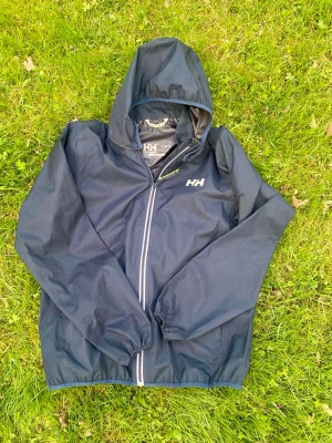 Svart regnjacka från Helly Hansen - Snygg marinblå regnjacka från Helly Hansen med huva och hel dragkedja framtill. Jackan har elastiska muddar och HH-logga på bröstet. Perfekt för regniga dagar och enkel att ta med sig. Materialet är vattenavvisande och lätt.