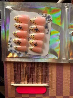 Press-on naglar - Snygga press-on naglar i mandelform med nude bas, gula spetsar dekorerade med svarta prickar och blommor. Inkluderar lim, fil och manikyrpinne. Perfekt för dig som vill ha en unik och trendig nageldesign.