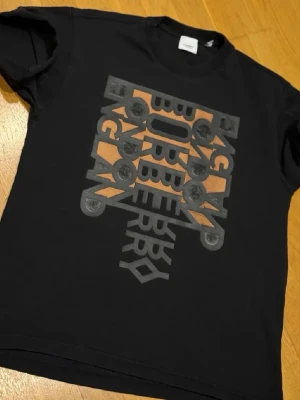 Svart Burberry t-shirt med tryck - Cool svart t-shirt från Burberry med grafiskt tryck i grått och orange på framsidan. Klassisk rund halsringning och korta ärmar. Perfekt för dig som vill ha en stilren men ändå unik look.