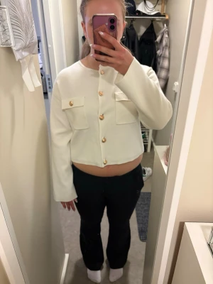 Vit cropped cardigan med guldknappar - Trendig vit cardigan med cropped passform och långa ärmar. Tröjan har två bröstfickor med lock och snygga guldiga knappar framtill. Perfekt för att lyfta en enkel outfit och ge en stilren look.
