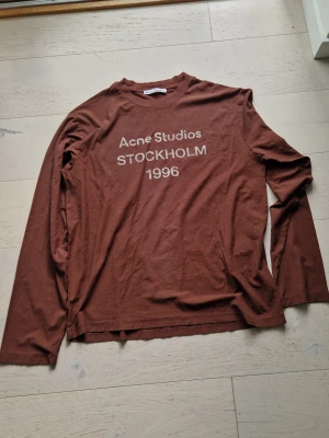 Brun långärmad topp Acne Studios L/M unisex - Säljer en brun långärmad topp från Acne Studios med tryck i vitt framtill där det står 'Acne Studios STOCKHOLM 1996'. Toppen har rund hals och är i mjukt bomullsmaterial. Perfekt för en avslappnad och trendig look.