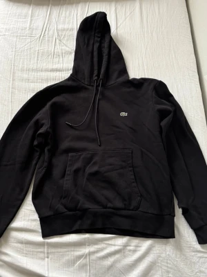 Svart hoodie från Lacoste - Svart hoodie från Lacoste med klassisk krokodillogga på bröstet. Hoodien har en stor huva med dragsko och en rymlig magficka. Tillverkad i mjukt material som känns skönt mot huden. Perfekt för en avslappnad och stilren look.