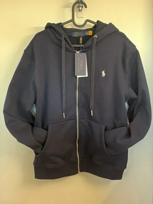 Polo Ralph Lauren Hoodie - Snygg mörkblå hoodie från Polo Ralph Lauren med dragkedja, två stora fickor fram och justerbar huva med snören. Klassisk logga broderad på bröstet. Tillverkad i mjuk bomullsblandning som känns riktigt skön mot huden.