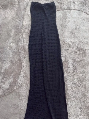 Svart lång stickad strandklänning - Svart långklänning i stickat material med snygg twistdetalj vid bysten. Klänningen är ärmlös och har ett hålmönster som ger en cool och trendig look. Perfekt för dig som gillar en stilren och elegant vibe på stranden.