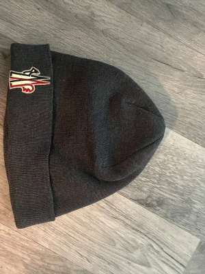 grå Moncler mössa med brodyr - Snygg svart stickad mössa från Moncler med uppvikt kant och broderad logga framtill i rött, vitt och beige. Klassisk modell som passar perfekt till kalla dagar. Materialet är mjukt och värmande, perfekt för vintern.