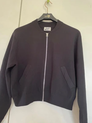 Svart bomberjacka från Acne Studios - Minimalistisk svart bomberjacka från Acne Studios med rak passform, silverfärgad dragkedja och två snedställda fickor framtill. Jackan har ribbade muddar vid hals och ärmslut och är tillverkad i ett mjukt tyg.