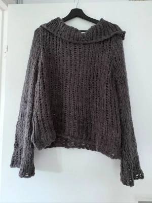 Grå oversize stickad tröja - Mjuk och luftig grå stickad tröja med oversize passform och vida ärmar. Tröjan har ett grovmaskigt mönster och en bred krage. Perfekt för lager-på-lager och chill dagar. Materialet ser ut att vara en fluffig ull- eller mohairblandning.