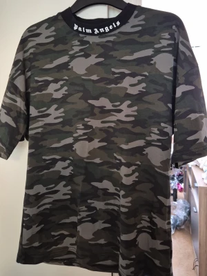 Palm Angels camo t-shirt - Snygg t-shirt från Palm Angels med klassiskt camouflage-mönster i gröna och svarta toner. T-shirten har korta ärmar och en svart krage med vit Palm Angels-logga. Perfekt för en streetwear-look.