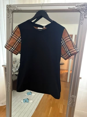 Svart burberry t-shirt med rutiga ärmar - Svart t-shirt med klassiska korta ärmar i beige och brunt rutig mönster. T-shirten har en rak passform och rund halsringning. Perfekt för en trendig och avslappnad stil. STORLEK L BARNSTORLEK