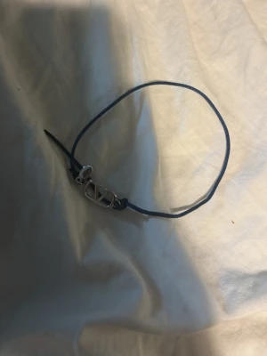 Valentino armband med blå snöre - Snyggt armband från Valentino med ett tunt blått snöre och en silverfärgad metallberlock med V-logga. Perfekt för dig som gillar stilrena accessoarer med designer-touch. Justerbar knut för enkel passform.