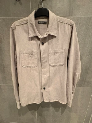 Beige overshirt från Dressmann - Stilren beige overshirt från Dressmann med två bröstfickor och knäppning framtill. Overshirten har klassisk krage och är tillverkad i ett mjukt, strukturerat tyg. Perfekt att slänga över en t-shirt för en avslappnad look.