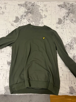 Olivgrön sweatshirt från Lyle & Scott - Olivgrön sweatshirt från Lyle & Scott med klassisk rund hals och lång ärm. Tröjan har en gul broderad logotyp på bröstet och ribbade muddar vid ärmslut och nederkant. Perfekt för en avslappnad och stilren look.
