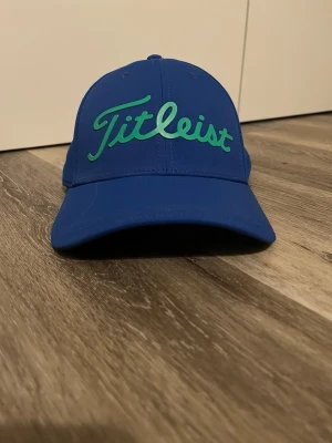 Blå Titleist keps med grön logga - Snygg blå keps från Titleist med böjd skärm och grön broderad logotyp framtill. Klassisk sexpanelsdesign med ventilationshål och justerbar passform. Perfekt accessoar för en sportig och stilren look. Pris kan absolut diskuteras.   