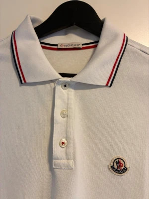 Vit pikétröja från Moncler - Klassisk vit pikétröja från Moncler med korta ärmar och krage. Tröjan har knappar framtill och Moncler-logga broderad på bröstet. Kragen och ärmsluten har röda och marinblå ränder för en sportig look.