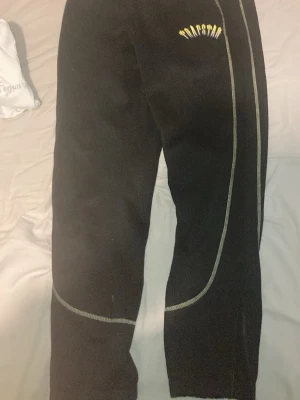 Svarta mjukisbyxor från Trapstar - Svarta mjukisbyxor från Trapstar med gul logga på ena benet och vita detaljer längs sidorna. Byxorna har dragsko i midjan och muddar vid bensluten. Perfekta för en avslappnad och trendig look.