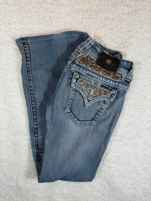 Miss Me ljusblå bootcut jeans storlek 27 - Snygga ljusblå bootcut jeans från Miss Me med dekorativa fickor bak, detaljer i silver och paljetter. Klassisk femficksmodell med låg midja och slitningar för en cool look. Perfekt för dig som gillar statement jeans.