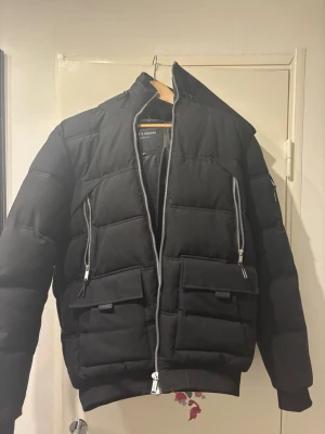 Supply and demand jacka - Snygg svart pufferjacka från Supply & Demand med två stora framfickor och två vertikala silverfärgade dragkedjor. Jackan har en boxig passform, ribbade muddar och hög krage. Perfekt för kalla dagar och ger en cool streetwear-look.