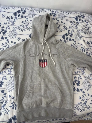 Grå hoodie från GANT med logga - Säljer en grå hoodie från GANT med broderad logga och emblem på bröstet. Hoodien har en stor ficka framtill, ribbade muddar och snören i huvan. Perfekt för dig som gillar klassisk och sportig stil. Materialet är mjukt och bekvämt.   OBS !! Finns en vit fläck tror det är slitet tror jag 