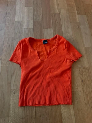Ribbad orange topp från Gina Tricot - Snygg ribbad topp i stark orange från Gina Tricot. Modellen är croppad med korta ärmar och har en unik v-ringad halsringning med liten slits framtill. Perfekt att styla med jeans eller kjol för en trendig look.