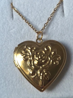 Guldfärgat medaljong hjärthalsband med ros i rostfritt stål  - Säljer ett guldfärgat hjärthalsband med vacker relief av en ros på framsidan. Halsbandet har en tunn kedja och hjärtat går att öppna för att lägga in en liten bild eller minne. Perfekt accessoar för dig som gillar romantiska detaljer och klassisk stil. Material: Rostfritt Stål (missfärgas inte). Kedjans längd: 45 cm + förlängningskedja. Hänget: Ca 3 cm