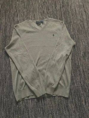 Grå  tröja från Polo Ralph Lauren - Snygg grå tröja från Polo Ralph Lauren med den klassiska broderade loggan på bröstet. Tröjan har långa ärmar och ribbade muddar vid ärmslut och nederkant. Perfekt att matcha med jeans eller chinos för en clean look.