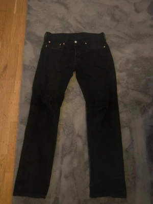 Svarta Levi's 501 jeans straight fit herrmodell - Klassiska svarta Levi's 501 jeans med rak passform och fem fickor. Jeansen har knappgylf och är tillverkade i slitstark denim. Perfekta för en avslappnad och tidlös stil. Storlek W32 L 34