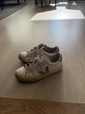 Vita Veja sneakers med beige detaljer - Snygga vita sneakers från Veja med beige detaljer och logga på sidan. Skorna har klassisk låg profil, vita snören och beige sula. Tillverkade i läder med textildetaljer på insidan. Perfekta för en clean och stilren look.