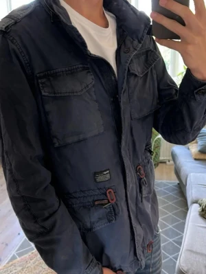 Mörkblå field jacket, size M - Snygg mörkblå field jacket med flera stora fickor framtill, axelklaffar och patch på ärmen. Jackan har dragkedja och detaljer i brunt samt en casual, normal passform. Perfekt för dig som gillar utility-stil och vill ha en jacka som sticker ut. Hör av dig vid minsta fundering och pris kan diskuteras! 🤩💯