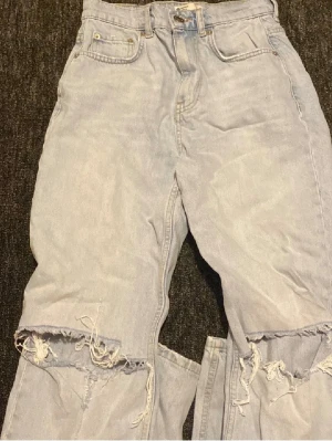 Ljusblå ripped jeans med hög midja - Säljer ett par ljusblå jeans med hög midja och slitna detaljer över knäna. Jeansen har klassisk femficksdesign och raka ben. Perfekta för en avslappnad och trendig look.