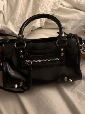 Svart handväska med silverdetaljer - Snygg svart handväska i glansigt konstläder med silverfärgade nitar och dragkedja framtill. Väskan har både handtag och avtagbar axelrem, samt coola metalldetaljer som ger en edgy vibe. Perfekt storlek för det viktigaste och passar till många olika looks.