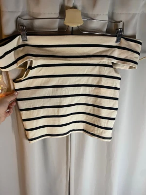 Randig offshoulder topp - Säljer en vit offshoulder topp med svarta horisontella ränder. Toppen är axelbandslös och har en croppad passform som ger en trendig look. Perfekt för dig som gillar en stilren och modern vibe.