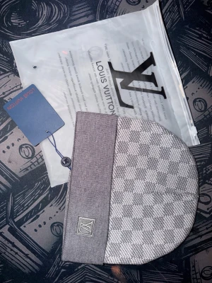 Grå Louis Vuitton Mössa - Snygg grå mössa från Louis Vuitton med klassiskt Damier-mönster i ljusgrå och mörkgrå nyanser. Mössan har en bred kant med LV-logga broderad framtill och är tillverkad i mjukt, stickat material. Perfekt accessoar för dig som gillar exklusiv och tidlös stil.