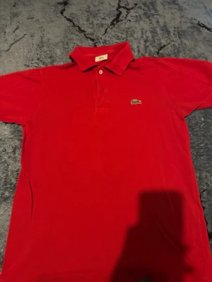 Röd pikétröja från Lacoste - Klassisk röd pikétröja från Lacoste med den ikoniska gröna krokodilloggan på bröstet. Tröjan har krage, knappar framtill och korta ärmar. Perfekt för en stilren och sportig look.