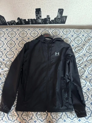 Svart vindjacka från Under Armour - Snygg svart vindjacka från Under Armour med huva och dragkedja framtill. Jackan har meshfoder för extra ventilation och två sidofickor. Perfekt för blåsiga dagar och sportiga outfits. Under Armour-logga på bröstet ger en sportig vibe.