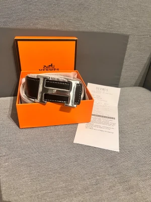  svart skärp med silver H - Snyggt svart skärp från H med stor, blank silverfärgad H-spänne. Skärpet är gjort i skinn och levereras i original orange H-box. Perfekt accessoar för att lyfta vilken outfit som helst.