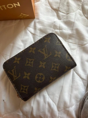 Louis Vuitton monogram plånbok - Säljer en brun plånbok från Louis Vuitton med klassiskt monogrammönster i guld. Plånboken har dragkedja i guldtonad metall och är tillverkad i tåligt canvasmaterial. Perfekt storlek för kort och sedlar, och en riktig ikon för dig som gillar lyxiga accessoarer.