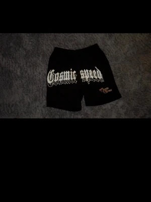 Svarta Cosmic Speed shorts - Svarta shorts från Cosmic Speed med stor vit text framtill och mindre logga i orange. De har en relaxed passform och resår i midjan för extra komfort. Perfekta för en avslappnad streetwear-look.