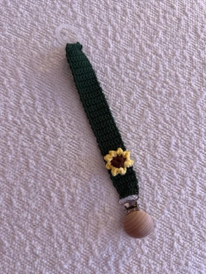 Grön virkad accessoar med solros - Handgjord virkad Napphållare i mörkgrönt garn med en dekorativ solros i gult och brunt. Utrustad med en träklämma och plastögla, perfekt som hållare. Materialet är garn och trä, och designen är unik och färgglad.